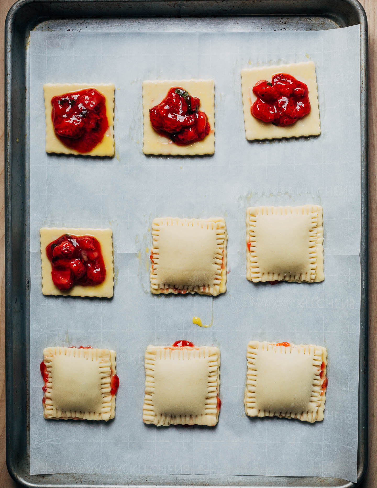Nine hand pies in the process of being assembled. Three pies have a bright red dollop of jam, and four others are covered and crimped.