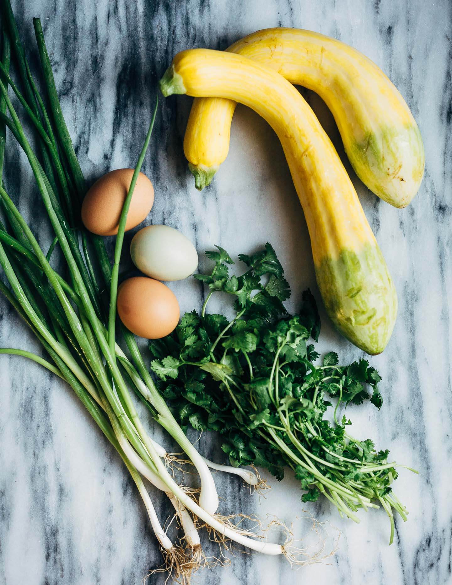 A marble slab with fritter ingredients: summer squash, eggs, green onions, and cilantro. 