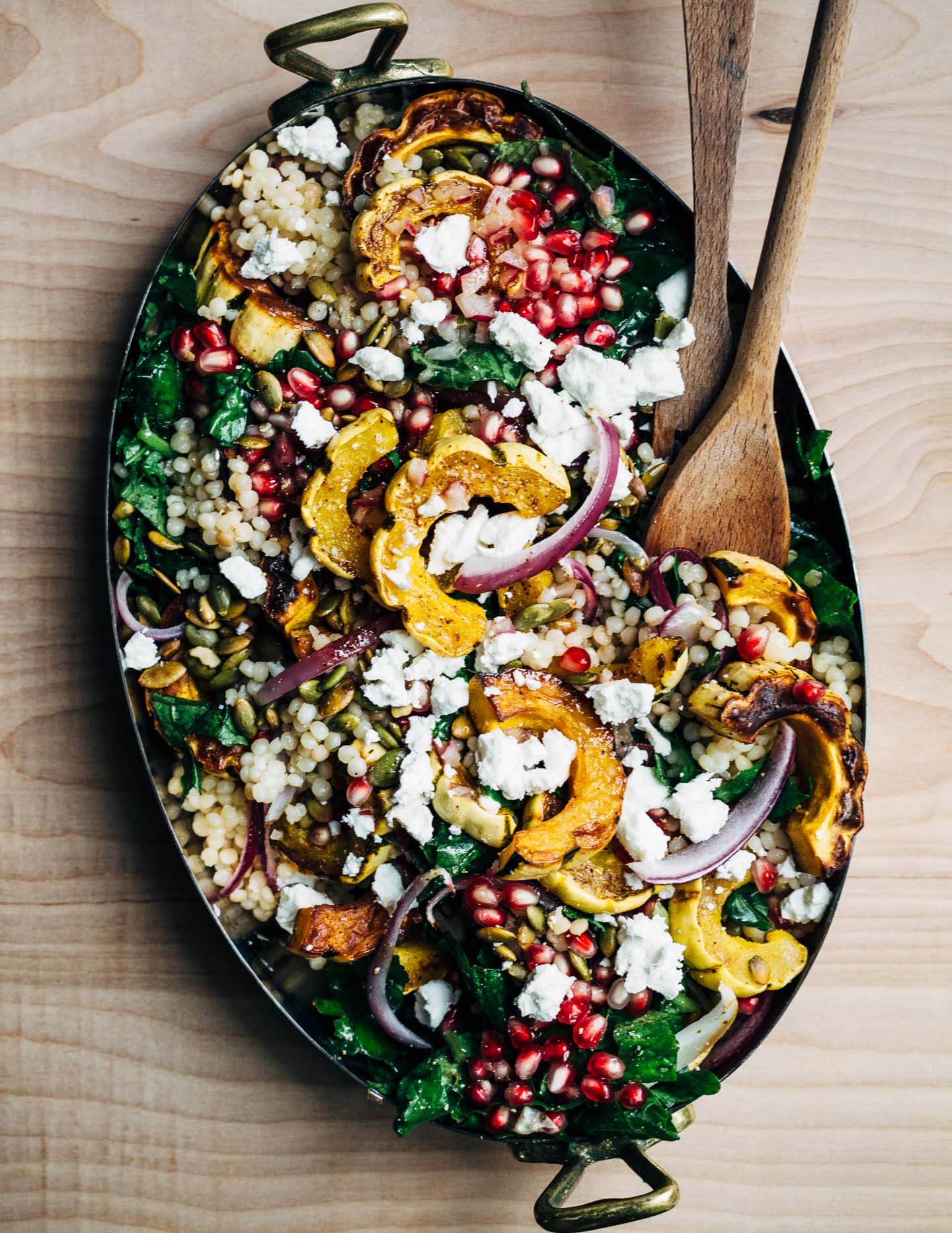 A serving dish filled with a couscous, delicata squash, and green salad. There are serving utensils in the salad. 