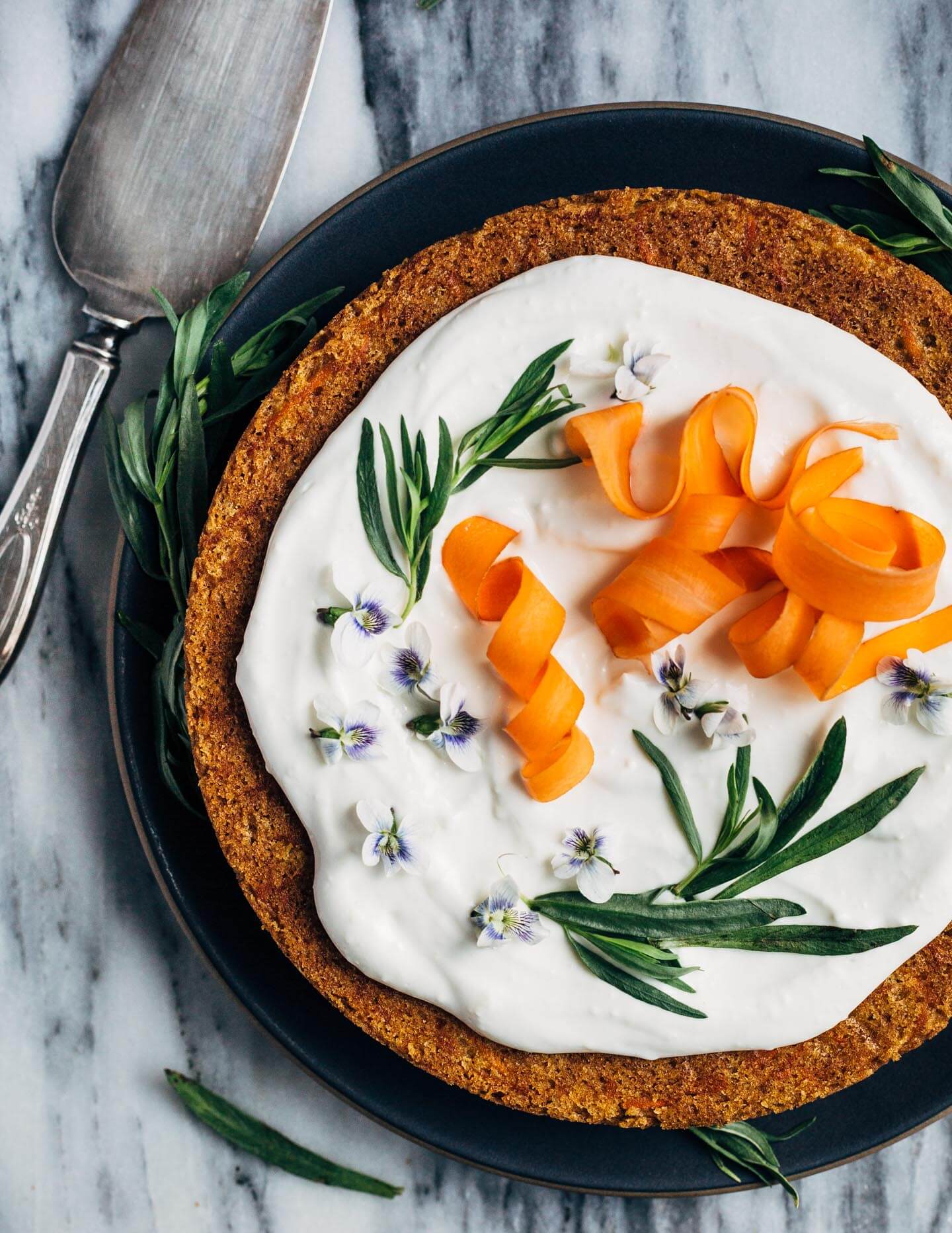 A simple olive oil carrot cake for spring celebrations! The tender, springy whole wheat crumb is suffused with nutty olive oil, fresh carrots, and orange zest. The cake is topped with an airy, lightly sweet yogurt and cream cheese frosting, carrot ribbons, tarragon, and edible flowers. 