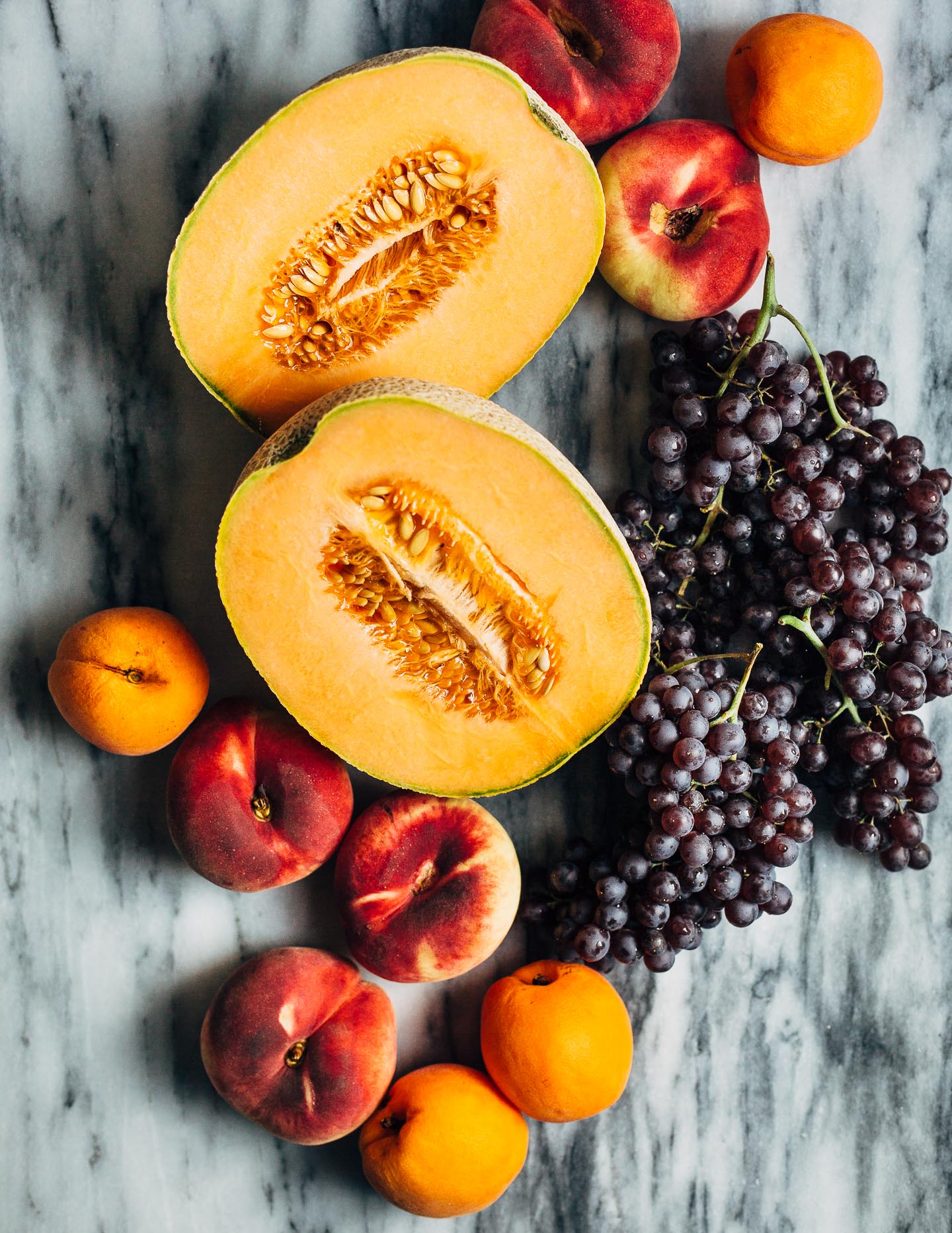 An array of fresh summer fruit: cantaloupe, yellow peaches, apricots, and Champagne grapes.