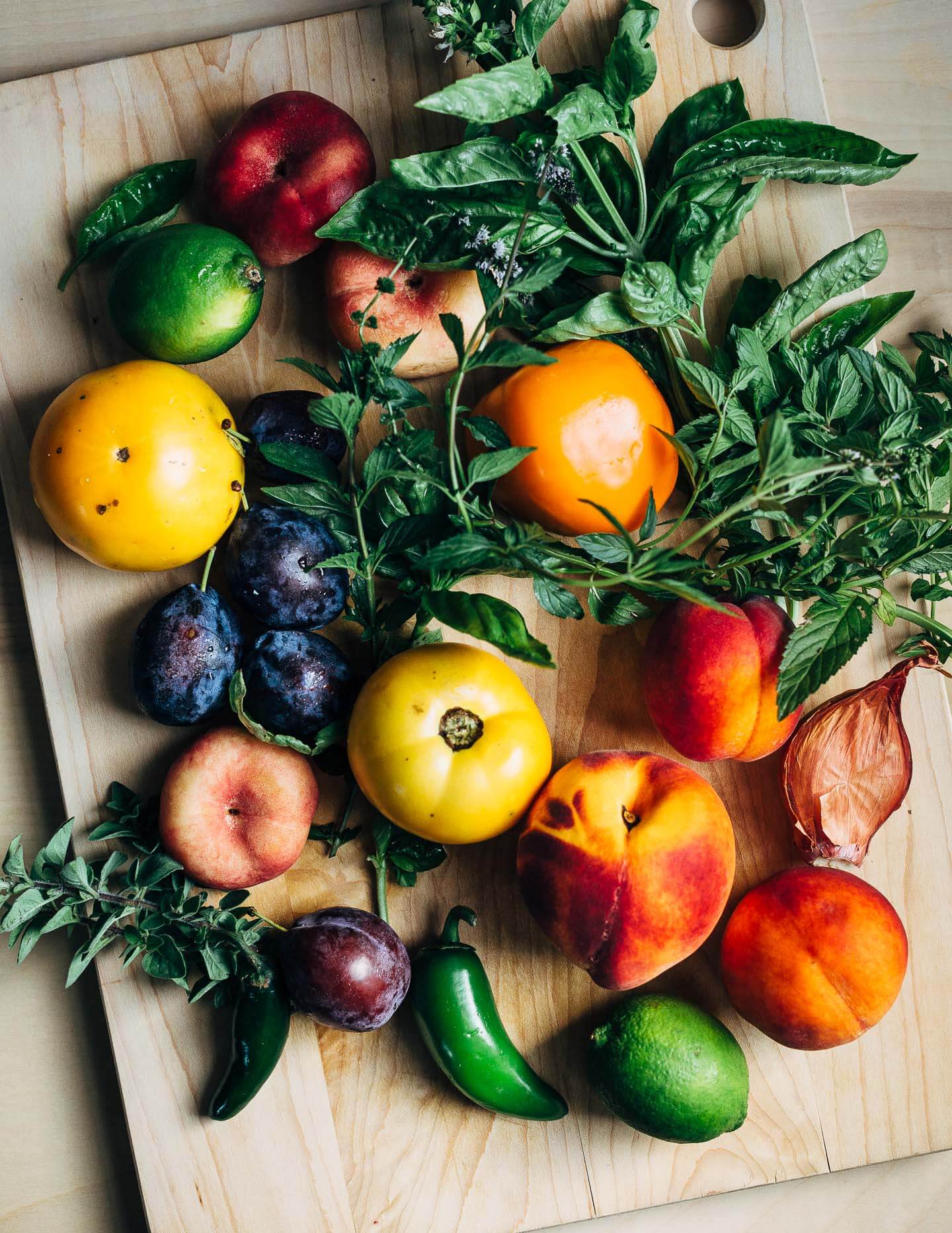 Late summer produce: yellow and donut peaches; damson plums; yellow, orange, and green tomatoes; fresh basil and mint.