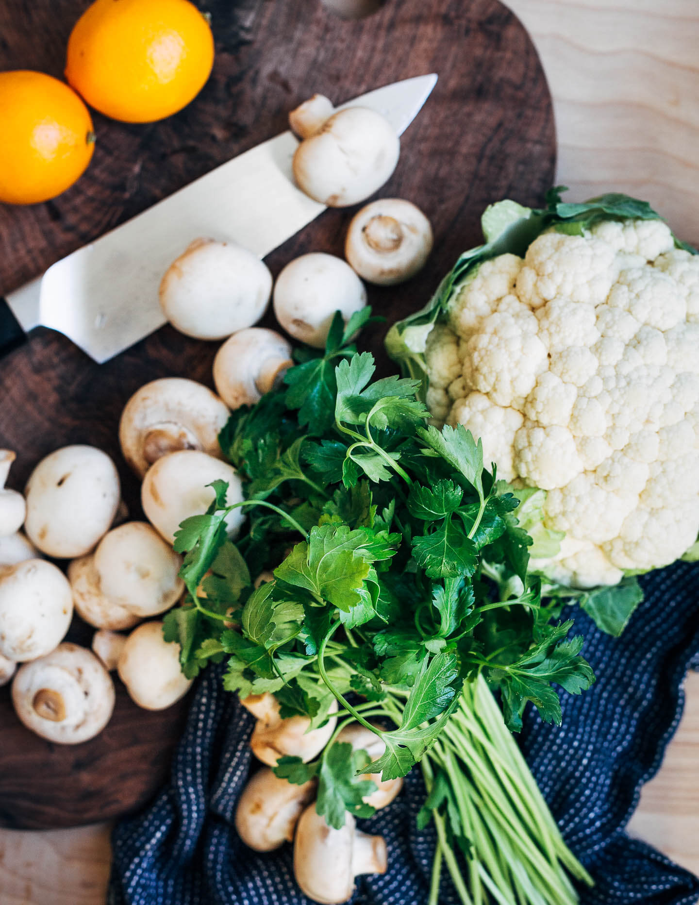Ingredients for roasted cauliflower and mushroom flatbreads. Ingredients include a whole head of cauliflower, a bunch of parsley, scattered button mushrooms, and two bright yellow Meyer lemons. 