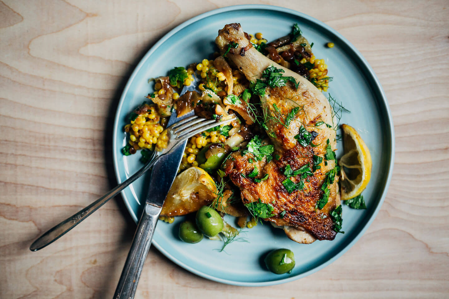 A small plate with a chicken legs, couscous, olives, and herbs.