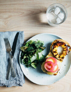 A dinner plate with an open bun. Topped with a grilled nectarine and a cheese-covered pork burger.