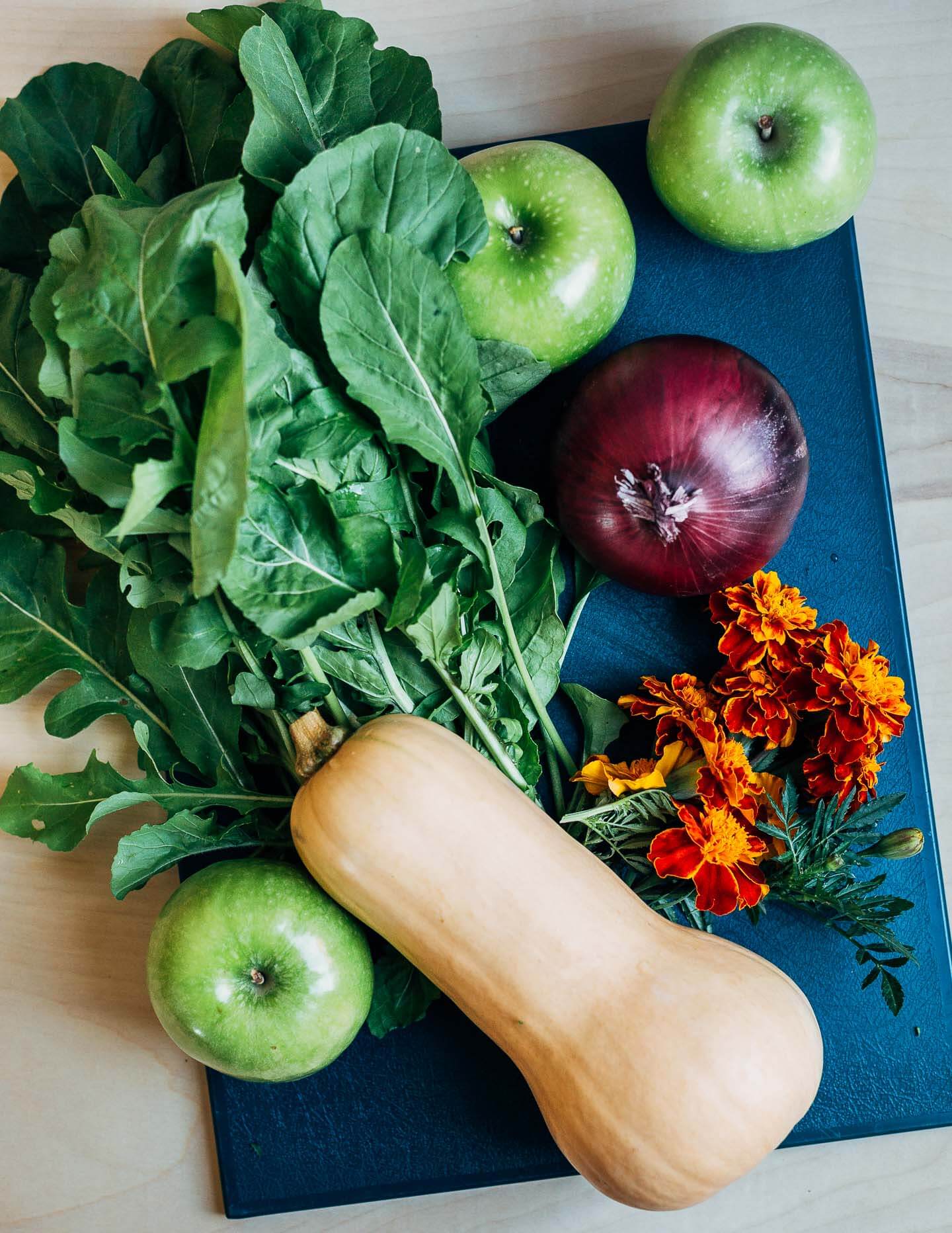 Arugula, butternut, onion, apples and marigolds arranged on a cutting board. 