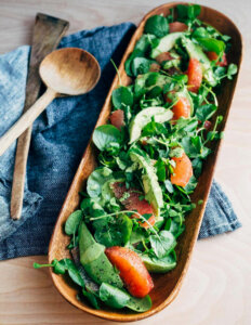A long, narrow wooden platter with baby green and sliced avocado and grapefruit.
