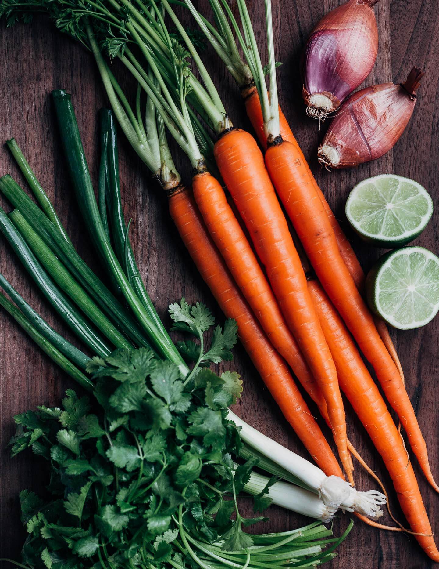 A cutting board with carrots, cilantro, green onions, shallots, and limes. 