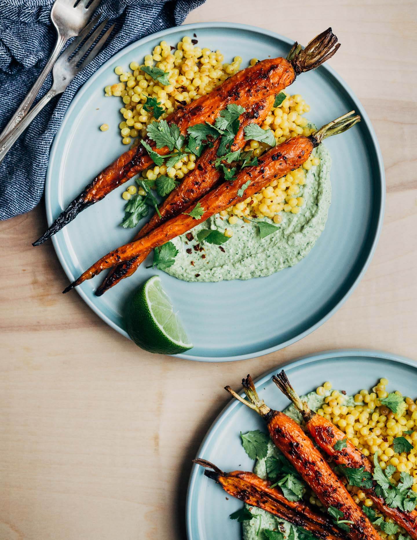 Two plates with roasted carrot, green tahini, and couscous. There are herbs and lime wedges scattered around the plates. 
