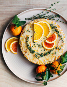 A whole cake decorated with orange slices and thyme sprigs on a pink platter.