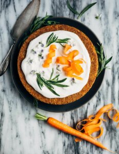 A carrot cake on a marble slab with frosting, carrot ribbons, and herbs on top.