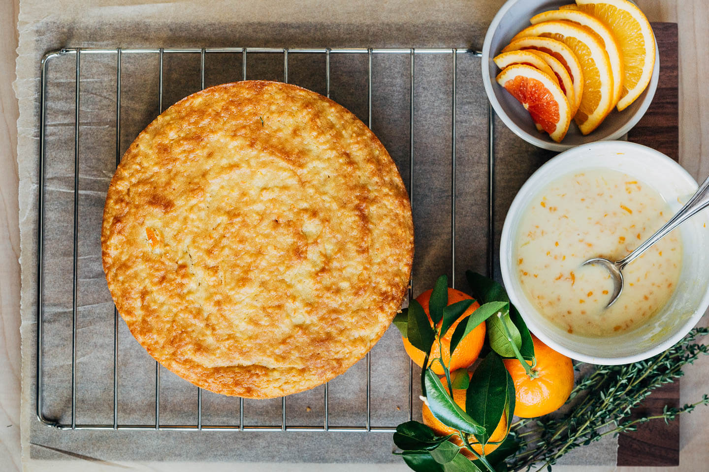 An orange cake on a wire rack, with a bowl of glaze, and sliced citrus alongside. 