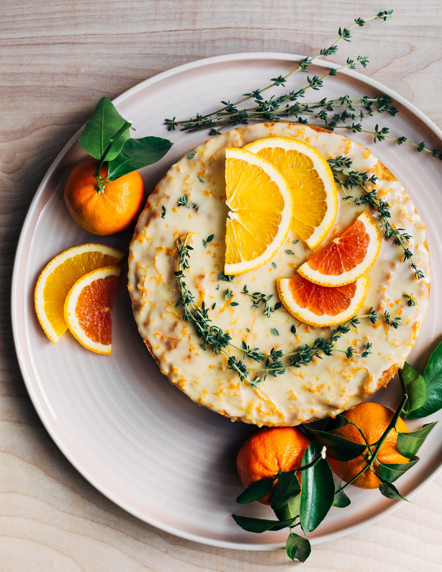 A platter with a glazed cake. It’s garnished with thyme sprigs, sliced citrus, and leafy whole citrus. 