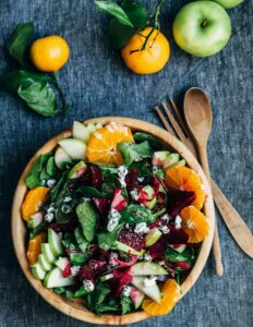 A wooden bowl with a salad made with apples, spinach, and oranges on a blue tablecloth.