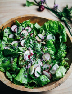 A wooden bowl with green leaves, sliced radishes, and herbs.