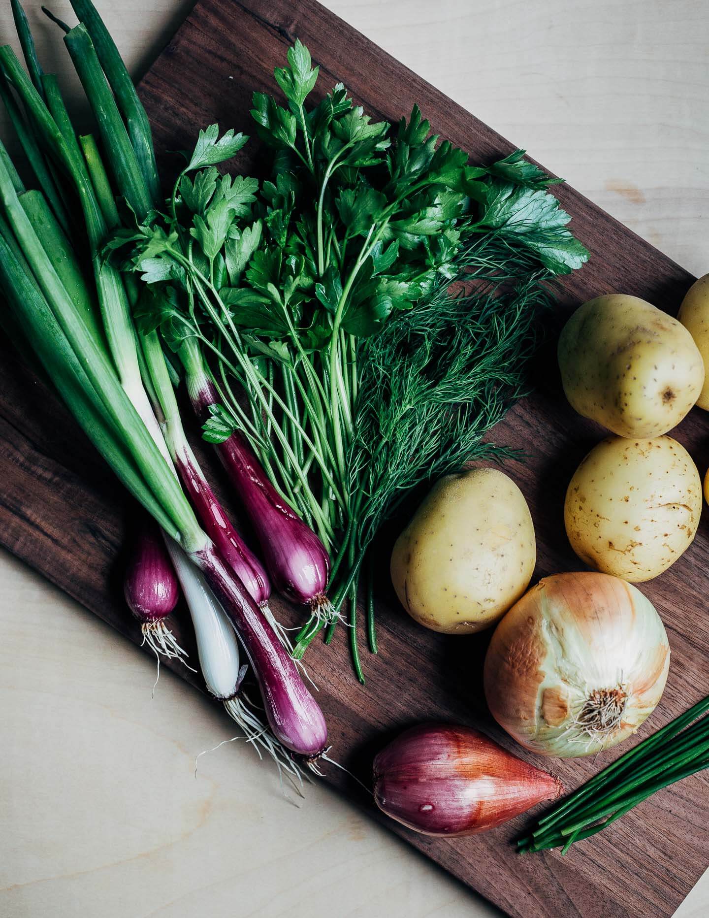 A cutting board with onions, potatoes, and herbs. 