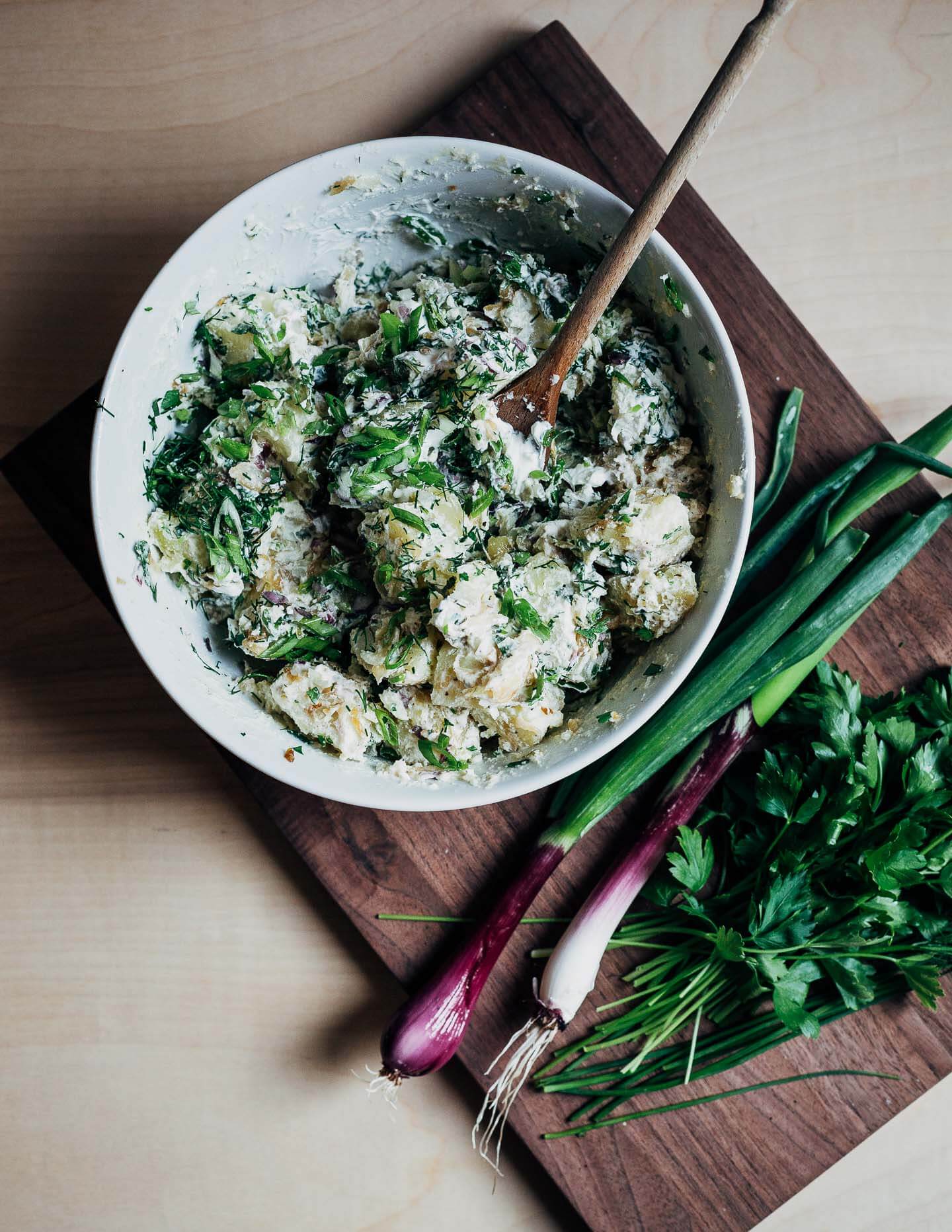 A mixing bowl of potatoes salad with spring onions and herbs on a cutting board alongside. 