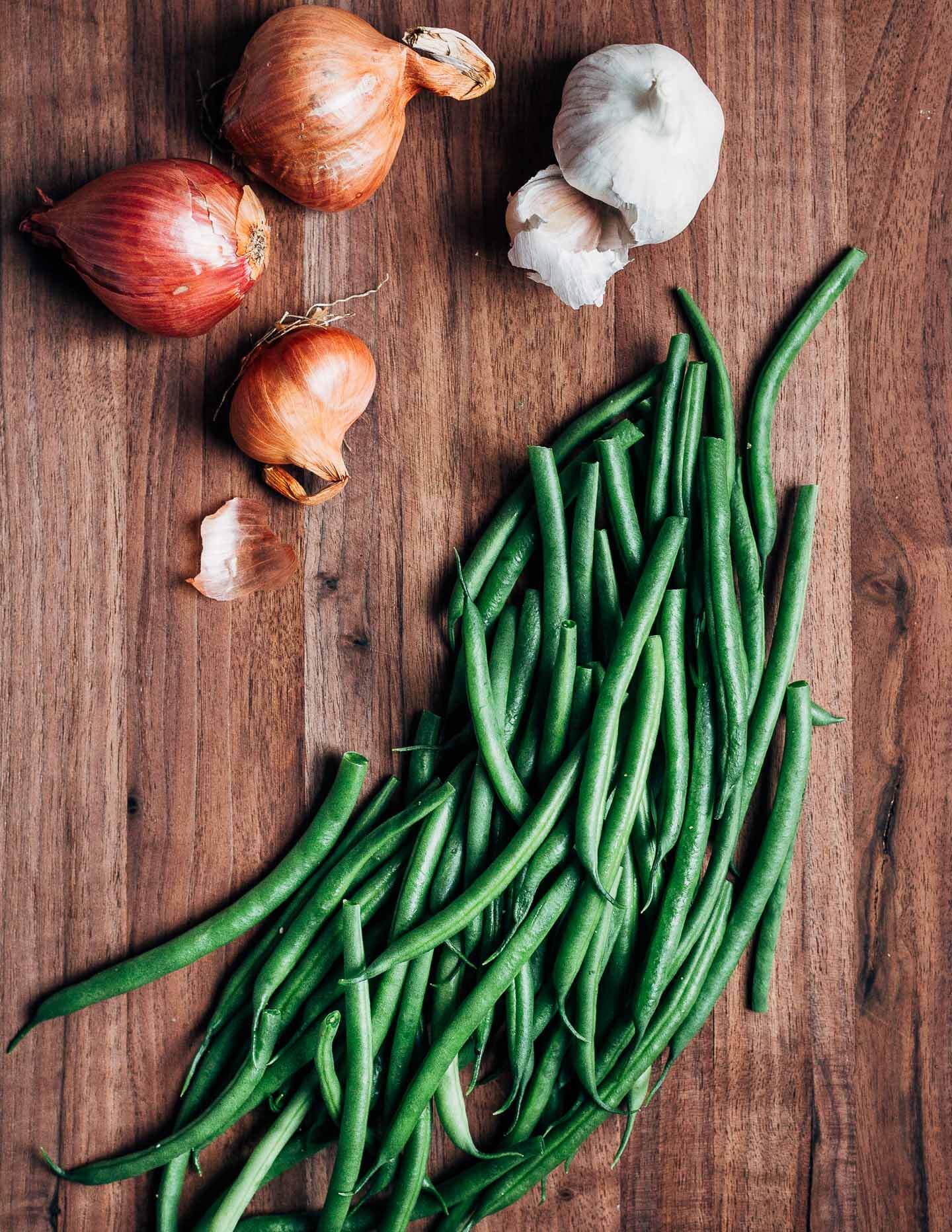 A dark wood cutting board with trimmed green beans, and shallot and garlic bulbs. 