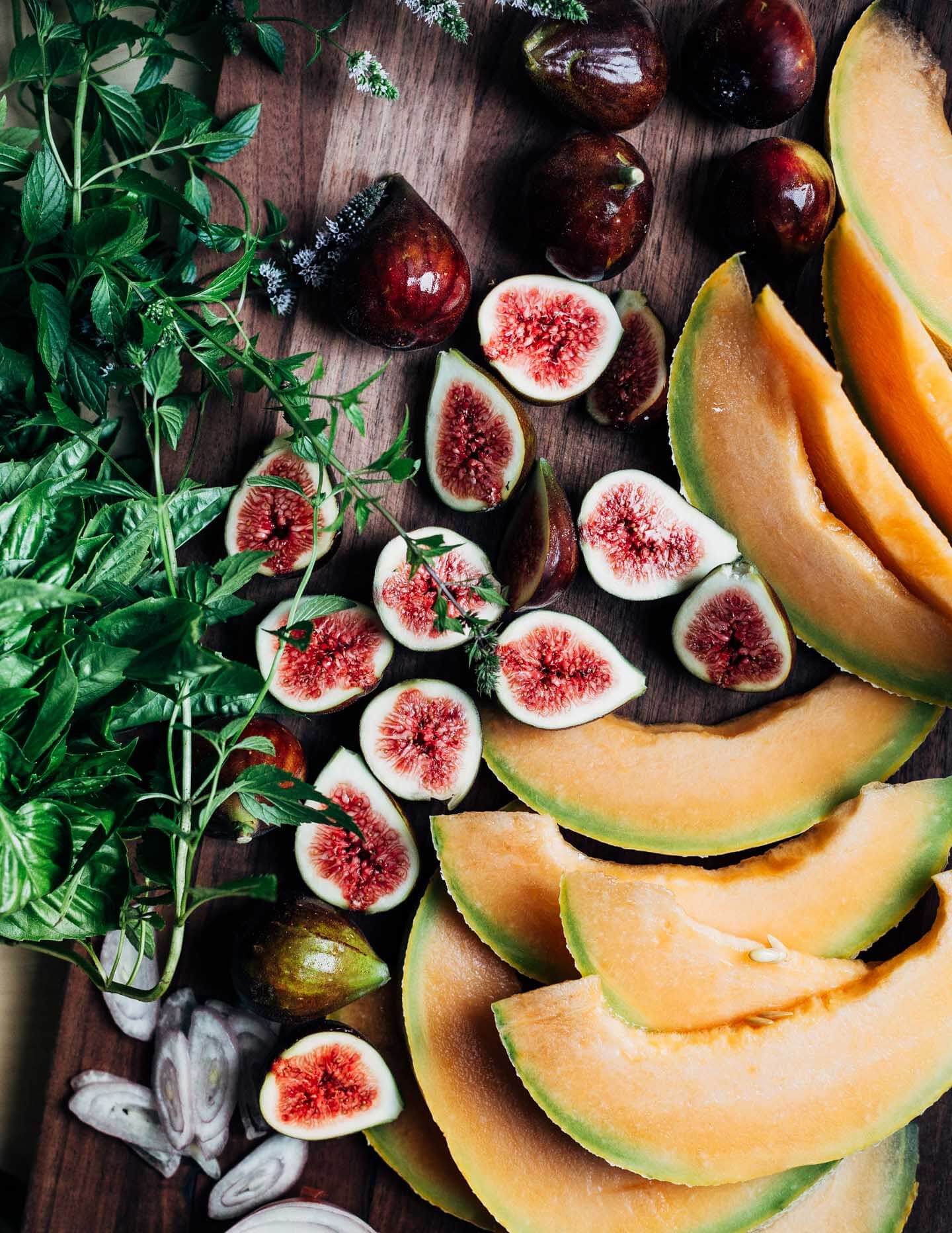 A cutting board with sliced cantaloupe, halved figs, and herbs.