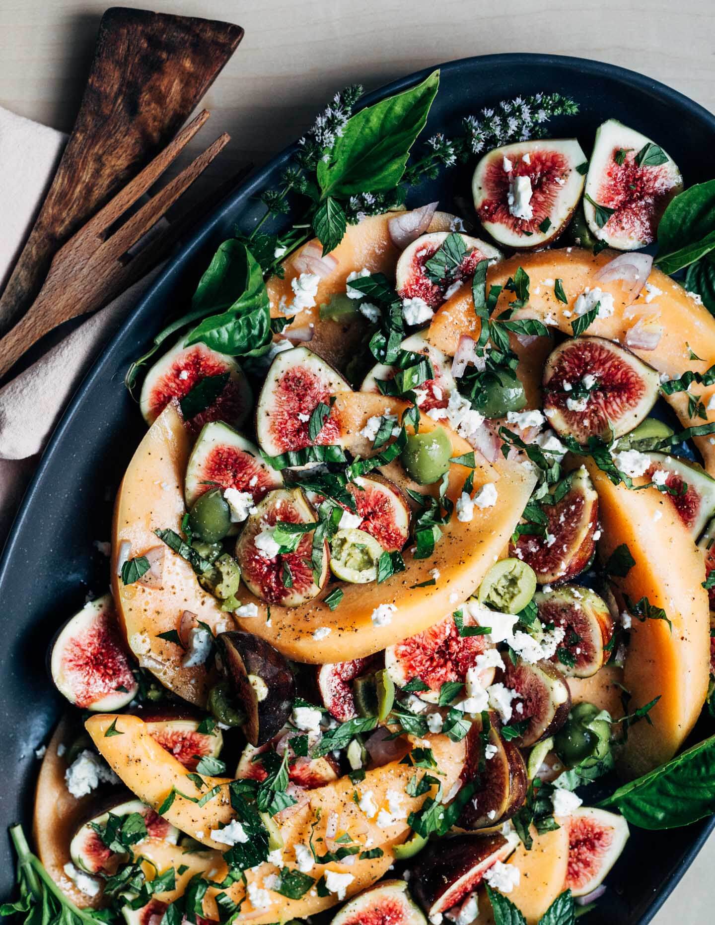 A large platter with cantaloupe, figs, feta, olives, and herbs. 