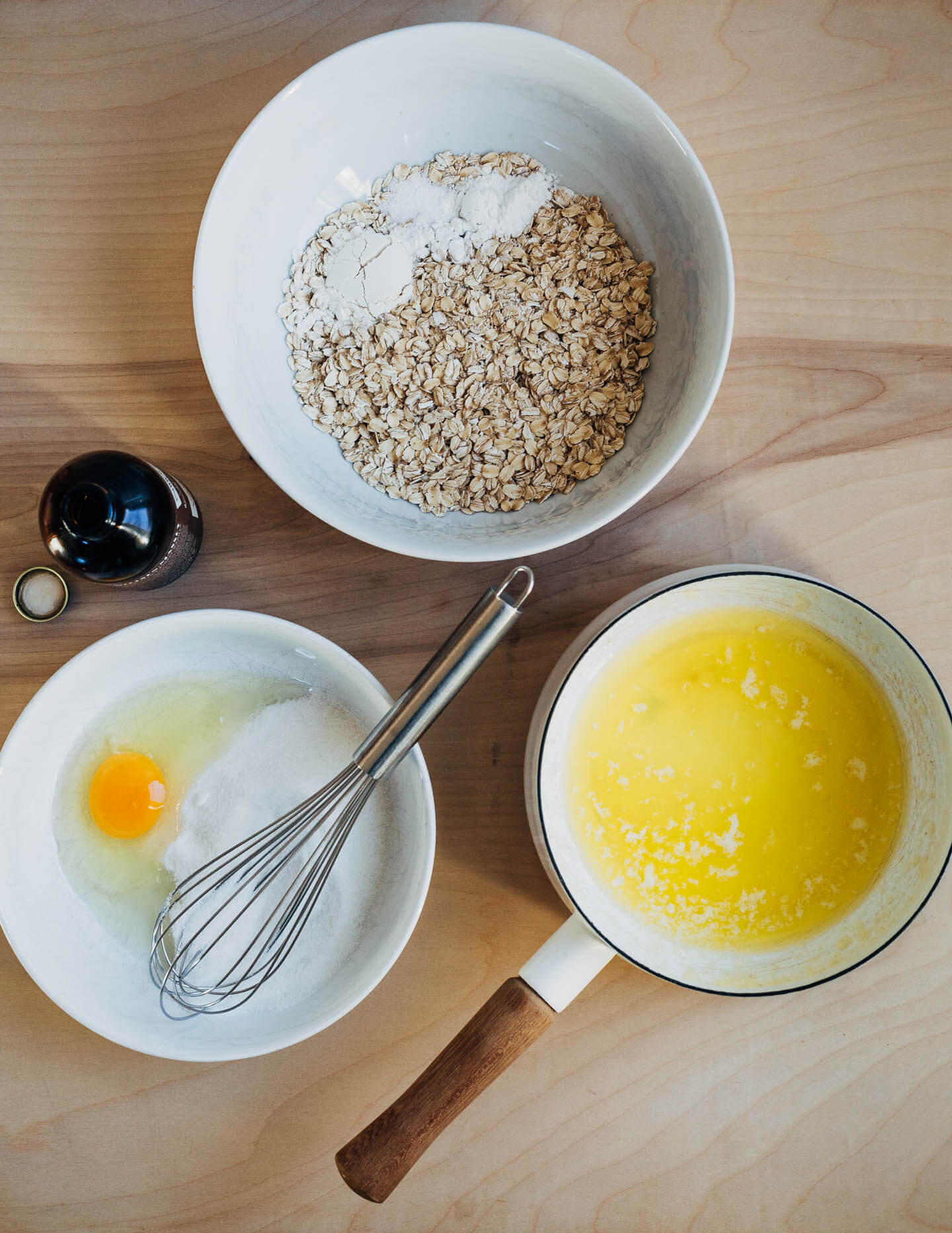 Ingredients for oatmeal cookies: melted butter in a saucepan, a bowl of oats, and a bowl with an egg, sugar, and a whisk. A bottle of vanilla extract sits alongside. 