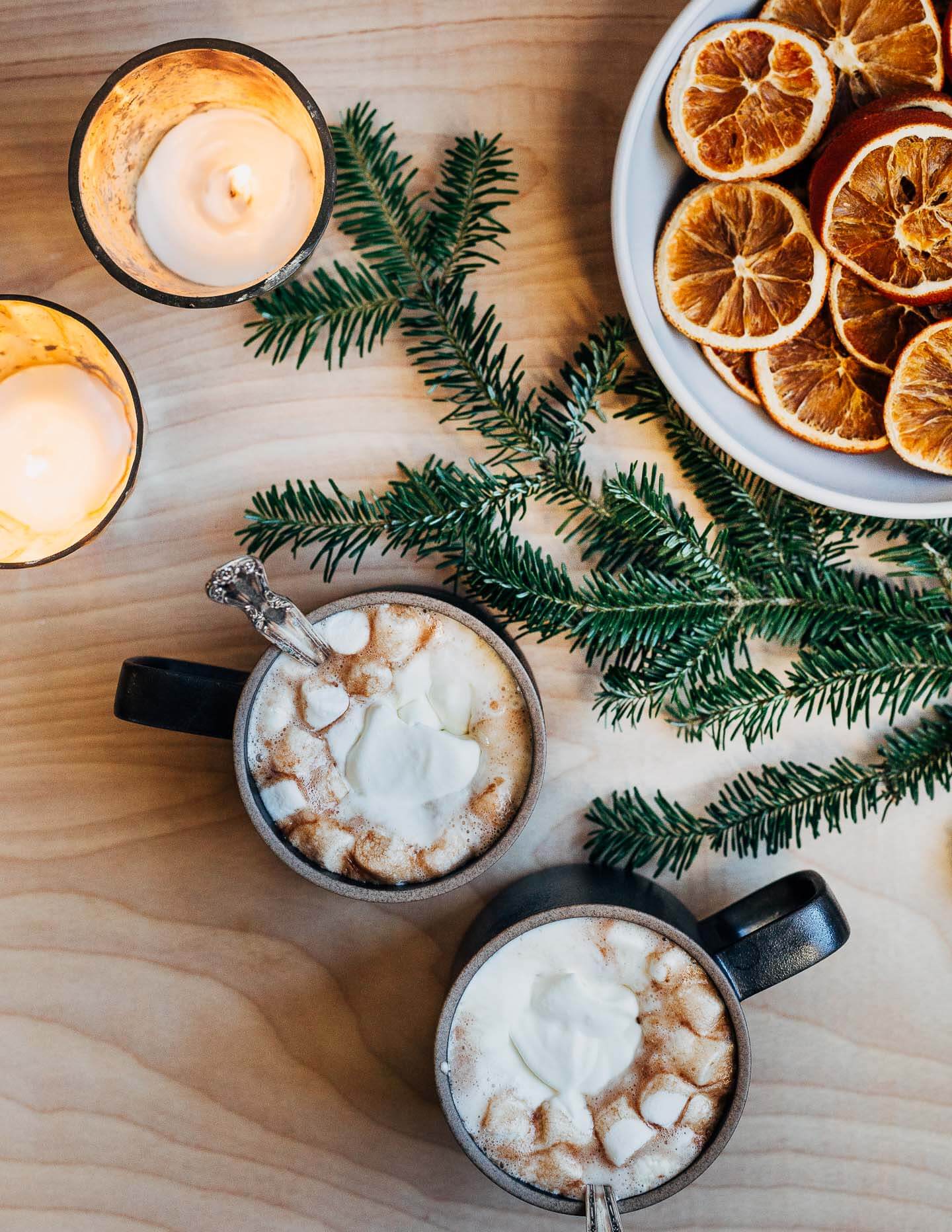 Two mugs of hot chocolate alongside candles, a pine bough, and a bowl of dried oranges. 