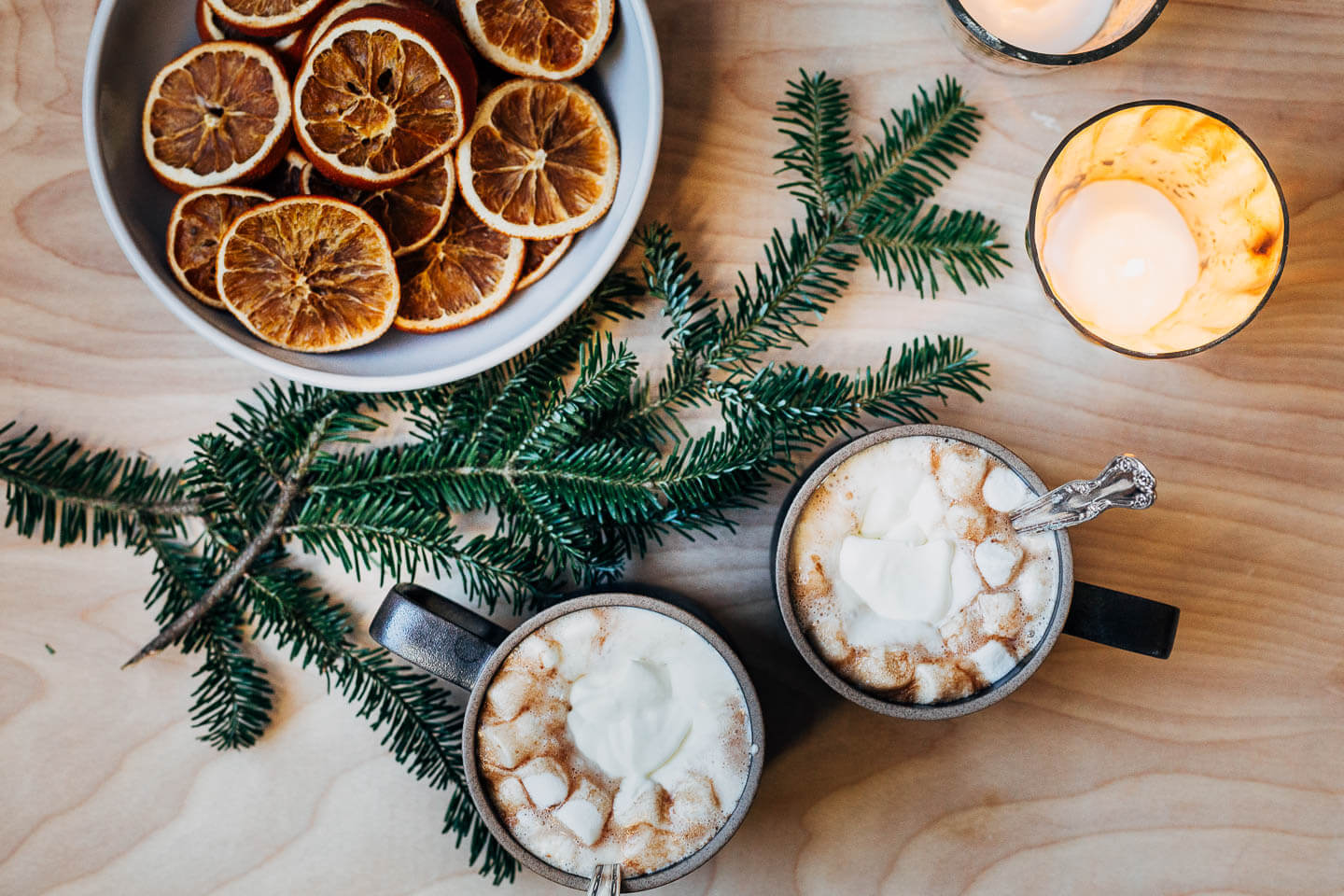 Two mugs of hot chocolate with candles, a pine bough, and a bowl of dried oranges.
