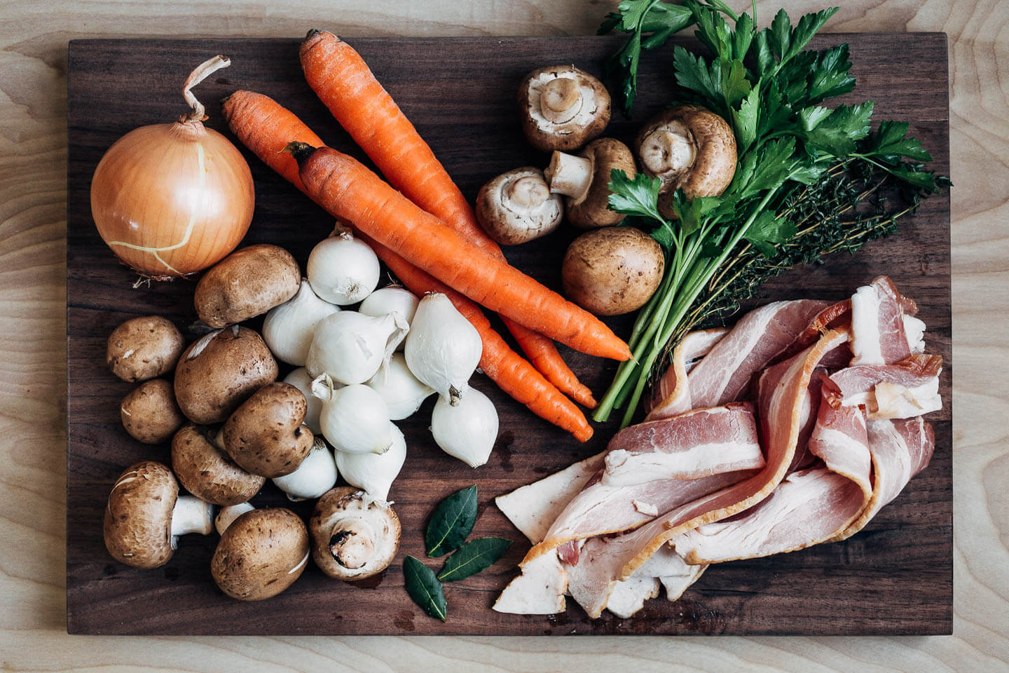 A cuuting board with onions, carrots, mushrooms, herbs, and bacon. 