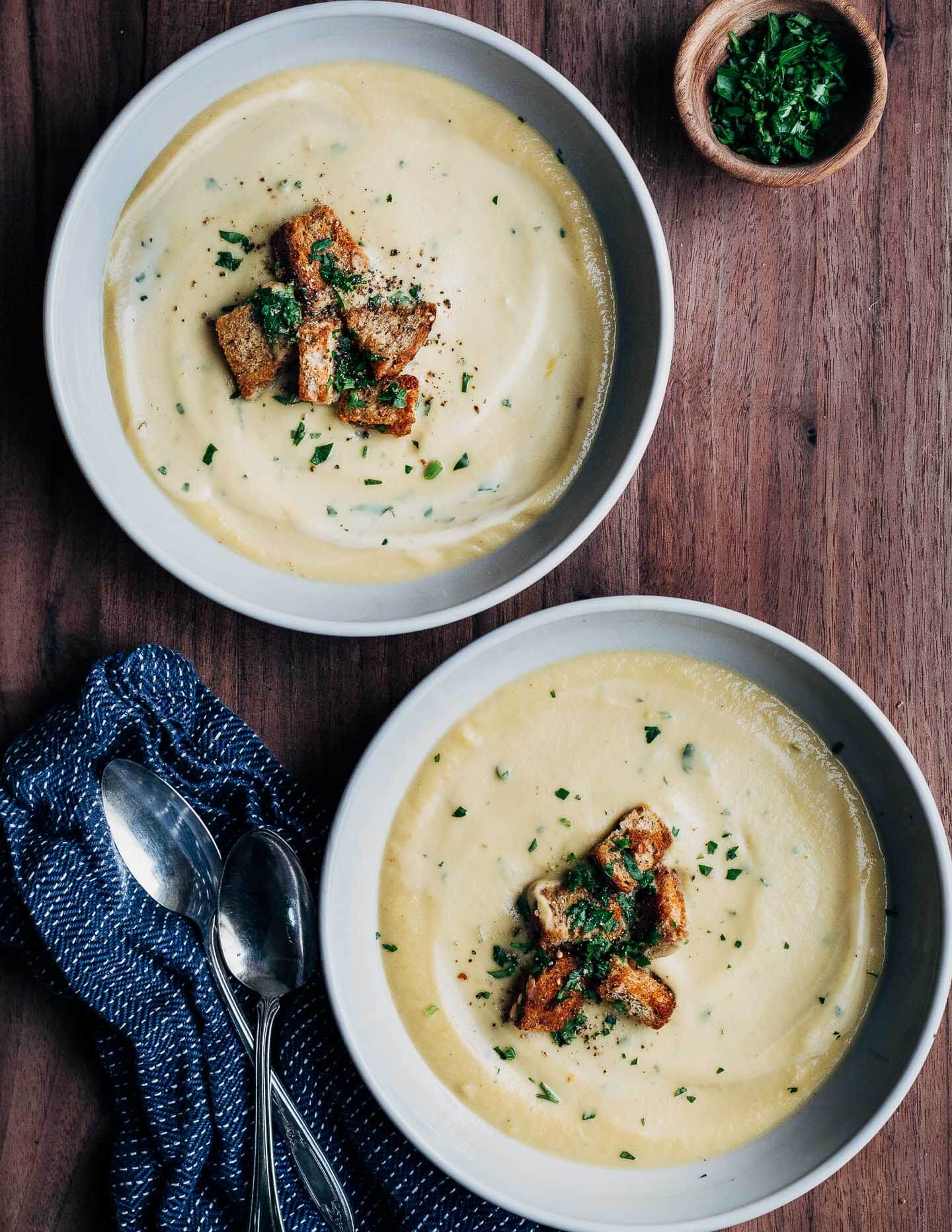 Two bowls of celery root soup on a dark wood table. There are napkins, spoons, and a bowl of minced alongside. 