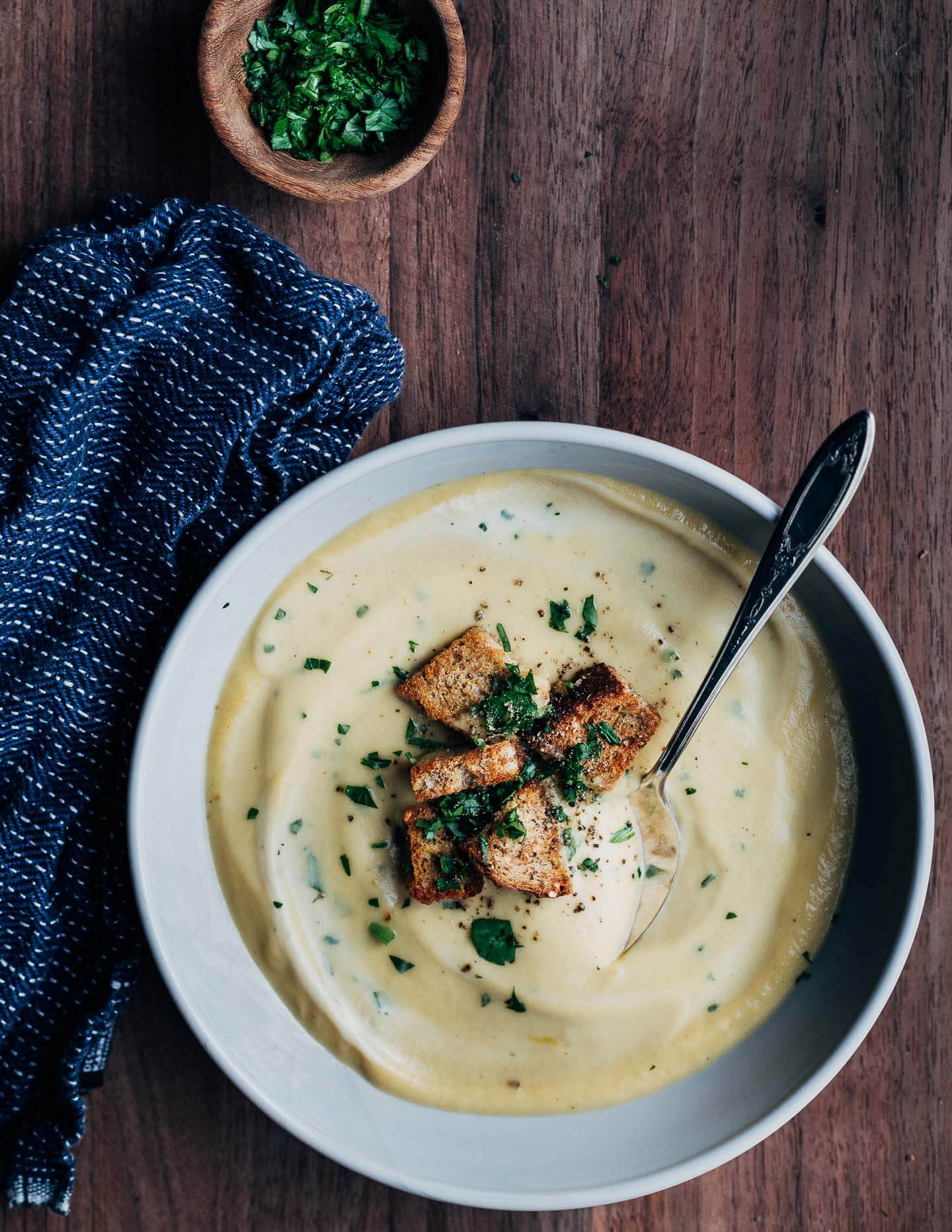 A spoon sitting in a bowl of celery root soup with a bowl of herbs and a blue napkin alongside. 