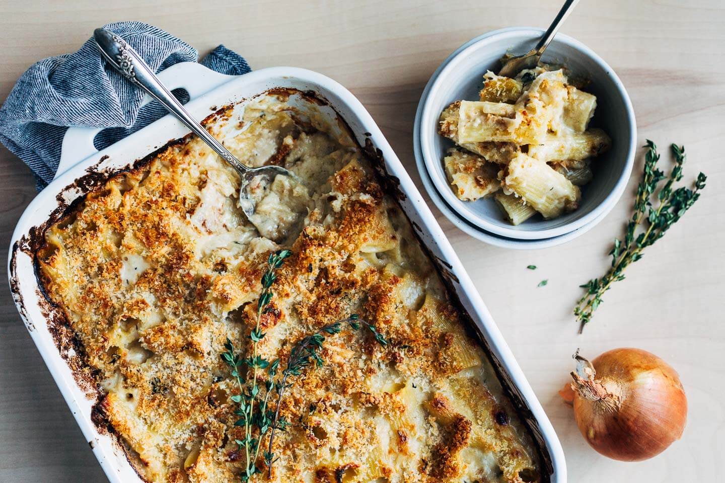 A baking dish with mac and cheese, with a serving spoon and a serving of mac in a bowl. There are herbs and an onion alongside. 