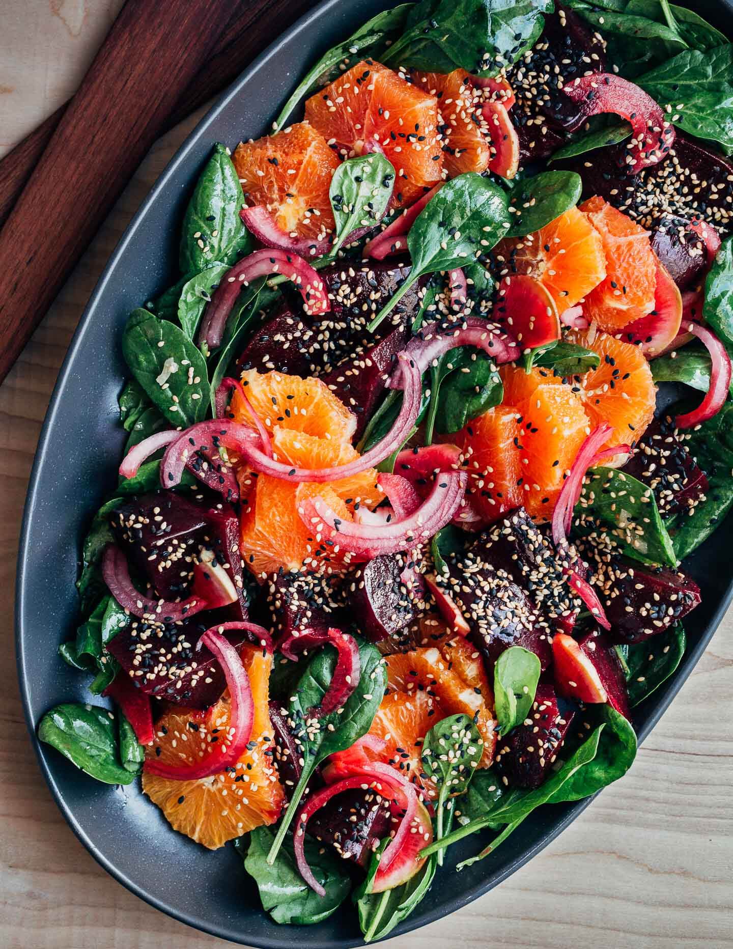 A platter with a colorful beet and citrus salad topped with sesame seeds. Salad tongs are alongside. 