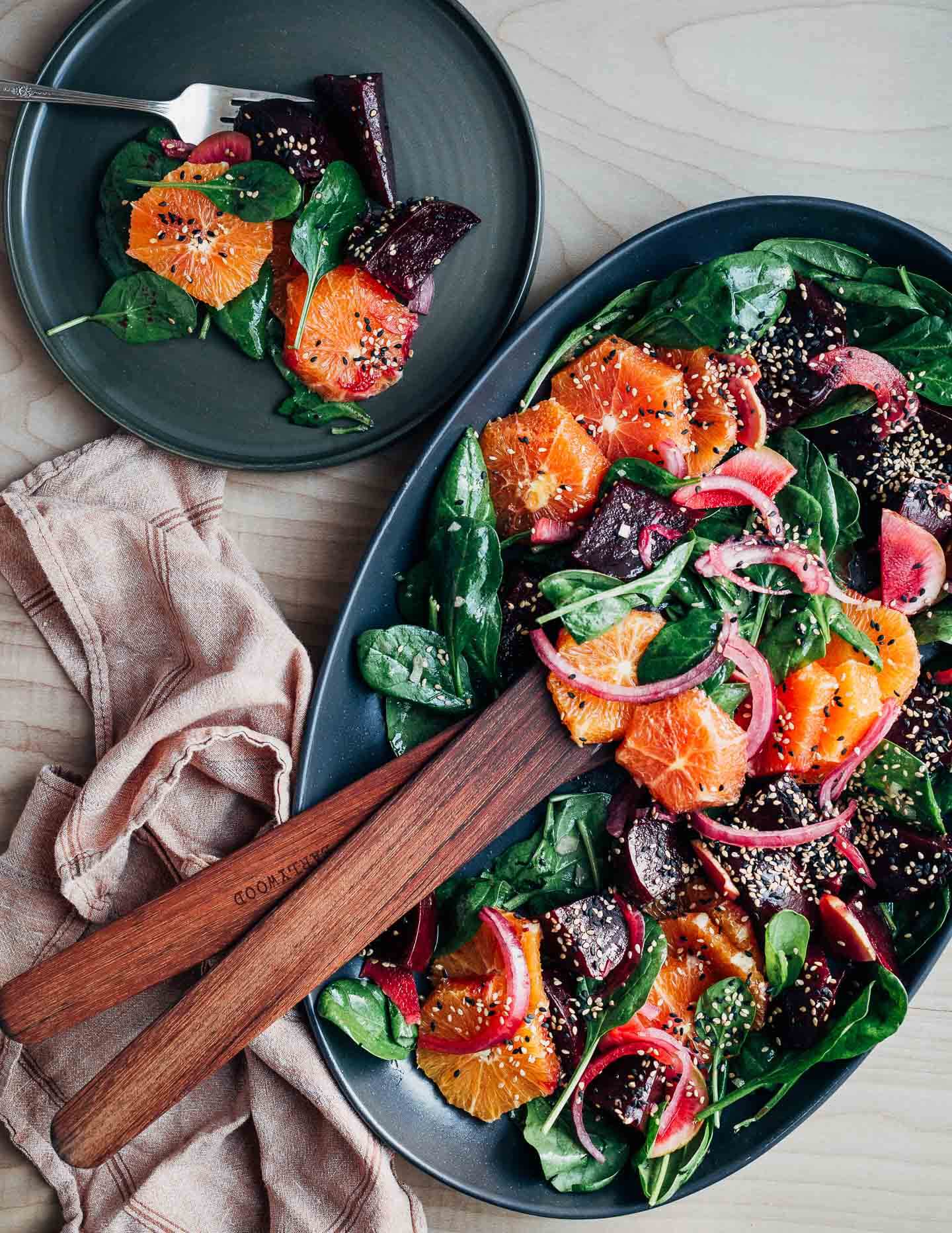 A platter with a colorful beet and citrus salad topped with sesame seeds. Salad tongs and a serving plate are alongside. 